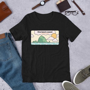 Peut inclure: T-shirt noir avec un graphique d'une île verte, un soleil et des nuages. Le texte "the best coast" et "Haystack Rock" est imprimé sur le graphique.