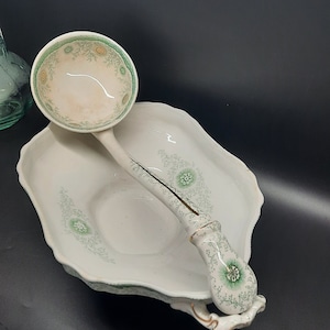 Puede incluir: Un tazón de servir de porcelana blanca con un patrón floral verde y una cuchara de servir a juego. El tazón tiene un borde festoneado y la cuchara tiene un mango decorativo.