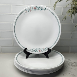 Könnte beinhalten: Ein Stapel weißer Keramikplatten mit grünem und floralem Rand. Eine Platte wird auf einem schwarzen Ständer präsentiert. Die Teller haben ein schlichtes, elegantes Design, das für den täglichen Gebrauch geeignet ist. Der Hintergrund zeigt eine weiße Fliesenspritzschutzwand.