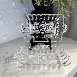 May include: Two clear glass serving dishes with scalloped edges and intricate floral patterns. The dishes are stacked on top of each other.