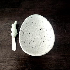 May include: An egg-shaped, speckled ceramic serving dish with a matching bunny-shaped spreader. The dish is light green with black speckles. The spreader has a light green bunny handle and a silver blade. The items are on a dark brown wooden surface.