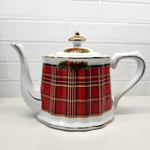 May include: A white porcelain teapot featuring a vibrant red, green, and gold tartan pattern around the body. The lid is topped with a gold-coloured finial, and the rim, spout, and handle are accented with gold trim. A festive holly and bow motif.