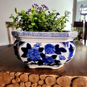 Könnte beinhalten: Ein dekorativer Keramik-Blumentopf mit blau-weißem Blumendesign. Der Topf ist mit grünen und lila Kunstpflanzen gefüllt. Der Topf steht auf einer Holzoberfläche.