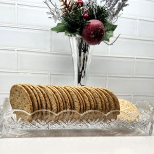 May include: A clear, decorative glass serving tray filled with round crackers. The tray has a scalloped edge and a starburst design. A vase with a holiday floral arrangement is in the background.