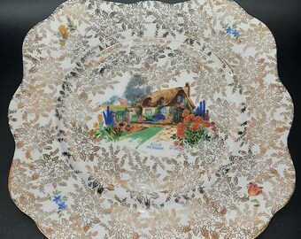 May include: A white ceramic plate with a gold floral pattern and a painted image of a cottage in a garden. The plate has a scalloped edge and says "A Glimpse of Old England" in the center.