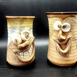 Peut inclure: Deux tasses en céramique faites à la main avec des visages fantaisistes. L'une des tasses a un large sourire avec des dents visibles et de grands yeux. L'autre tasse a une expression plus subtile. Les deux tasses sont de couleur beige avec des accents marron et blancs.