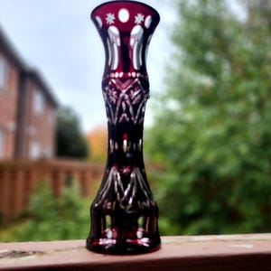 May include: A tall, slender cranberry red glass vase featuring intricate clear cut-glass patterns. The design includes geometric cross-hatching, star motifs, and vertical panels, with a flared rim and a wide, stable base for display.