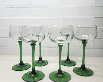 May include: A set of six clear glass goblets with deep green stems and circular bases. Each glass features a wide, rounded bowl atop a slender, colored stem that transitions into a solid green base, creating a uniform look for the collection.