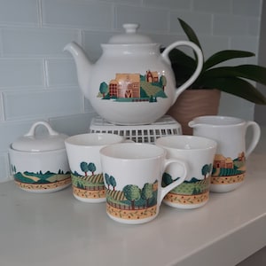 May include: A white teapot and four mugs with a country scene design. The teapot has a handle and a spout. The mugs have handles and are decorated with a green, brown, and yellow design of a farmhouse and trees.