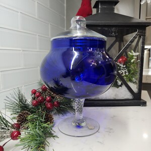 Peut inclure: Un grand bocal d'apothicaire en verre bleu cobalt avec un couvercle en verre transparent et une tige torsadée décorative. Le bocal repose sur une base circulaire transparente. Une lanterne noire et de la verdure de fête en arrière-plan.