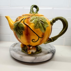 Può includere: Una teiera in ceramica a forma di zucca con finitura arancione lucida, caratterizzata da foglie e viticci verdi scolpiti. Il manico e il beccuccio sono verdi e una piccola figura di coniglio marrone è posizionata alla base della zucca.