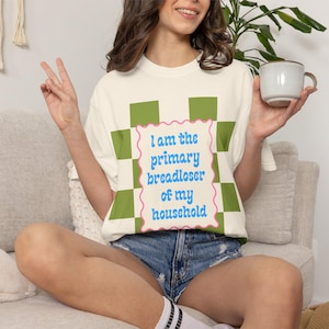 May include: Cream-colored t-shirt with the text "I am the primary breadloser of my household" in blue script. The text is framed by a pink wavy border and a green and white checkered pattern. The person is wearing denim shorts.