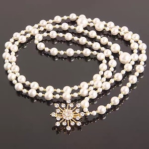 May include: A long white pearl necklace with a gold snowflake pendant. The necklace is made of multiple strands of pearls and is draped over a black surface.
