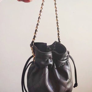 Op de afbeelding: Een zwarte quilted bucket bag met een gouden kettingband en een trekkoord.