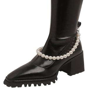 May include: Black knee-high boot with a square toe and a chunky heel. A white pearl chain is draped around the ankle. The boot has a lug sole.