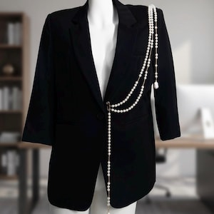 May include: A black blazer with a three-quarter sleeve design, paired with a layered pearl necklace. The necklace is draped over the blazer, adding a touch of elegance. The blazer is displayed on a mannequin.