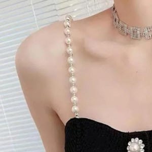 May include: A white pearl strap with a silver chain. The strap is worn over a black dress with a pearl button.