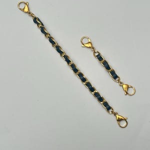 Op de afbeelding: Twee gouden kettingriemen met zwarte lederen accenten. De riemen hebben aan beide uiteinden kreeftklemmen.