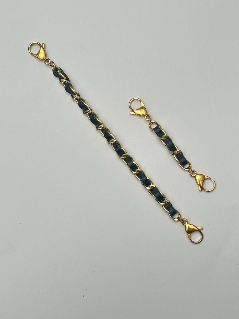 Op de afbeelding: Twee gouden kettingriemen met zwarte lederen accenten. De riemen hebben aan beide uiteinden kreeftklemmen.