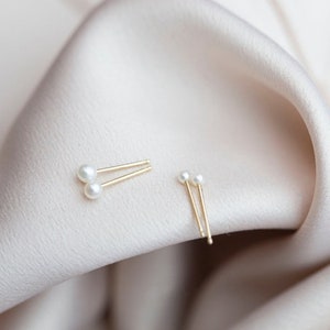 May include: A pair of gold stud earrings with small white pearl accents. The earrings are displayed on a soft, light pink fabric background.