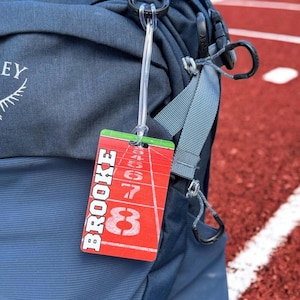 Könnte beinhalten: Ein rotes und grünes Gepäckanhänger mit dem Namen "BROOKE" in weiß auf rotem Hintergrund. Der Anhänger zeigt eine weiße Nummer "8" und andere Zahlen auf einer roten Bahn.