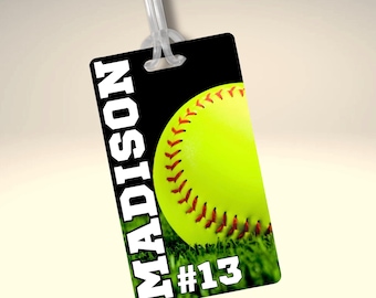 Könnte beinhalten: Ein schwarz-weißes Gepäckanhänger mit dem Namen "MADISON" und der Nummer "#13" in weißer Schrift. Ein gelber Softball mit roten Nähten auf grünem Gras.