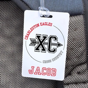 May include: White luggage tag with the text "Charleston Eagles" and "XC Cross Country" in black and red. The name "Jacob" is printed in red at the bottom. The tag is attached to a backpack.
