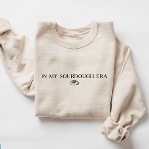 May include: A beige sweatshirt with the text "IN MY SOURDOUGH ERA" and a small illustration of a loaf of bread.