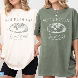 May include: Two women wearing oversized t-shirts with a vintage-style graphic design. The design features a sourdough bread loaf with the text "SOURDOUGH Social Club KELLY'S KITCHEN" printed in white on a green background.
