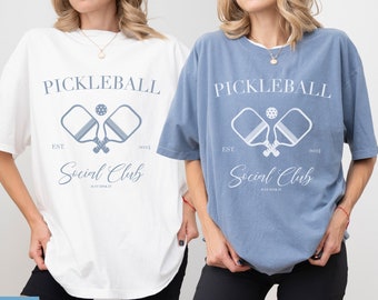May include: Two women wearing oversized t-shirts with a white graphic design that says "Pickleball Social Club" with a blue pickleball paddle and ball illustration. The t-shirt on the left is white and the t-shirt on the right is blue.
