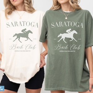 May include: Two short-sleeve crewneck t-shirts, one in ivory and one in moss green. Each shirt features a graphic of a jockey on a horse with the text SARATOGA, EST 2024, Bach Club, and SOPHIA'S BACHELORETTE printed in a mix of serif and script fonts.