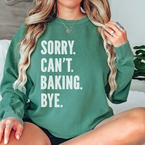May include: A light green sweatshirt with the text "SORRY. CAN'T. BAKING. BYE." printed in white.