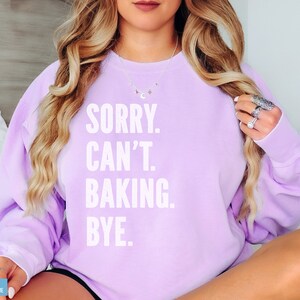 May include: A lavender sweatshirt with the text "SORRY. CAN'T. BAKING. BYE." printed in white.