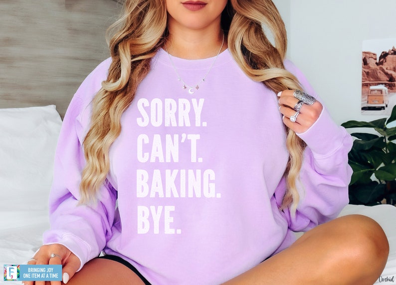 May include: A lavender sweatshirt with the text "SORRY. CAN'T. BAKING. BYE." printed in white.