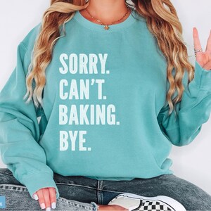May include: A teal blue sweatshirt with the text "SORRY. CAN'T. BAKING. BYE." in white.