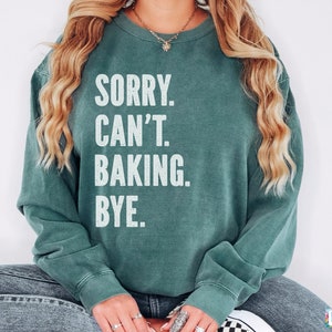 May include: A teal green sweatshirt with the text "SORRY. CAN'T. BAKING. BYE." printed in white.