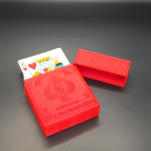 May include: A red Bicycle Standard playing card case with a matching lid. The case is open, revealing a playing card with the King of Hearts. The case has the Bicycle logo and spade symbol.