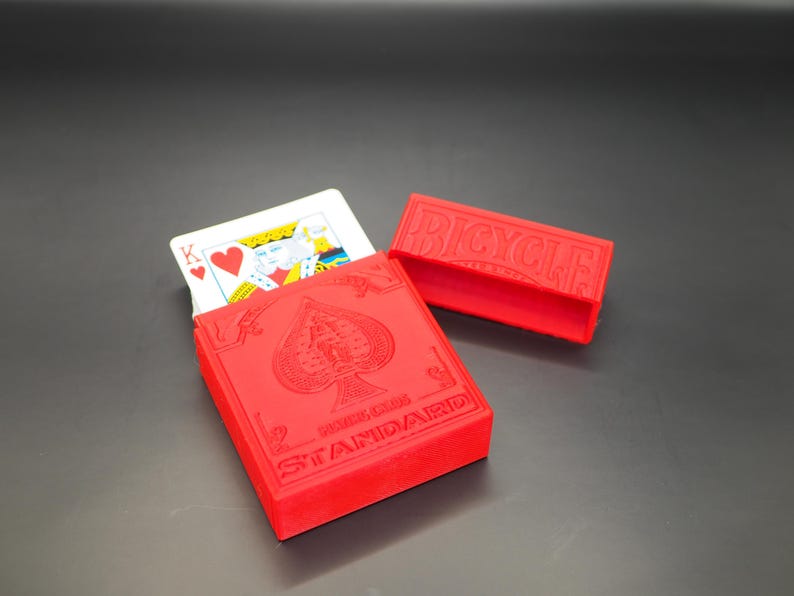 May include: A red Bicycle Standard playing card case with a matching lid. The case is open, revealing a playing card with the King of Hearts. The case has the Bicycle logo and spade symbol.