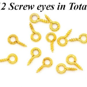 May include: Twelve gold screw eyes, also known as screw-in eyelets, are scattered on a white surface.  These are useful for attaching jewelry, leather, or other crafts.