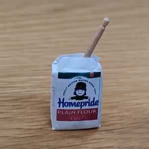May include: A white bag of Homepride Plain Flour with a red and green label. The bag is 1.5kg and features a cartoon chef with a red hat and a blue shirt. A wooden stick is sticking out of the top of the bag.