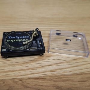 May include: A miniature black Technics turntable with a gold-coloured arm and a clear plastic cover. The turntable has the Technics logo in white and green. The turntable is on a wooden surface.