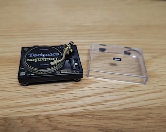 May include: A miniature black Technics turntable with gold-colored arm and a clear plastic cover. The turntable has the Technics logo in white and green. The turntable is on a wooden surface.