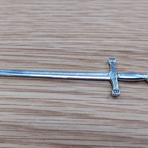 May include: A detailed, silver miniature sword with an ornate hilt and a pointed blade. The hilt features decorative carvings and a circular emblem at the pommel. The sword is displayed on a light brown wooden surface.