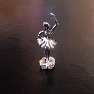 May include: A small, silver-colored ballerina figurine in a graceful pose. The dancer is wearing a tutu and has one arm raised. The figurine is mounted on a clear, textured base. The background is a dark, reflective surface.