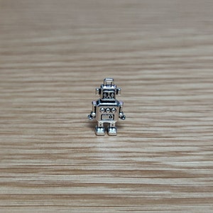 May include: Silver robot charm with a round head and four limbs. The robot has a small antenna on top of its head and a small circle on its chest.