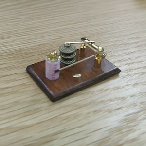 May include: A miniature wooden base with a gold metal winding mechanism and a pink and gold cylinder. The mechanism has a small wooden handle and a gold metal rod.