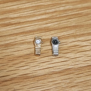 May include: Two miniature wristwatches. One is gold-toned with a white watch face and a silver-toned band. The other is silver-toned with a black watch face and a silver-toned band. Both watches are on a wooden surface.