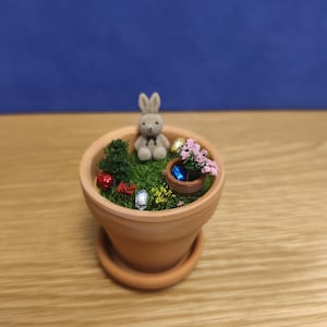 May include: A miniature Easter garden scene in a terracotta pot. The scene includes a small gray bunny, miniature Easter eggs, a small green tree, and pink flowers. The pot sits on a matching saucer, creating a charming, festive display.