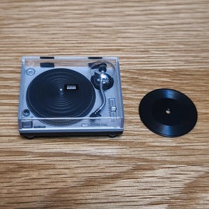 May include: A miniature silver Technics turntable with a black record and a clear plastic cover. The turntable has a silver tone arm and a black platter. The record is black and sits next to the turntable. The item is on a wooden surface.