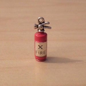 May include: A miniature red fire extinguisher charm with silver-colored details. The charm has a small label with the word "FIRE" and an "X" symbol. The charm is a small, detailed replica of a fire extinguisher.